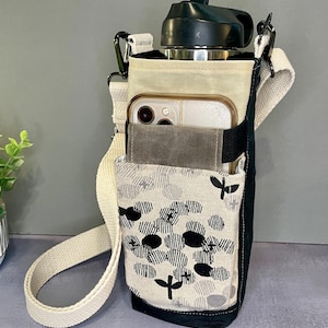 May include: A black and cream fabric water bottle carrier with an adjustable off-white shoulder strap. The carrier features a front pocket containing a smartphone and a secondary pocket with a black and gray circular pattern on a cream background.