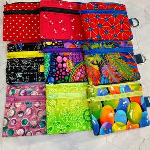 May include: A collection of small, zippered pouches in various colorful patterns. Designs include red polka dots, abstract shapes, feathers, buttons, and balloons. Each pouch has a contrasting zipper and a small metal ring.