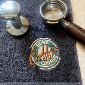May include: A dark gray towel with a green and white embroidered logo that says "Rob's Coffee Corner". The logo has a brown outline and features a stylized image of a coffee cup. A silver portafilter and tamper are also on the towel.