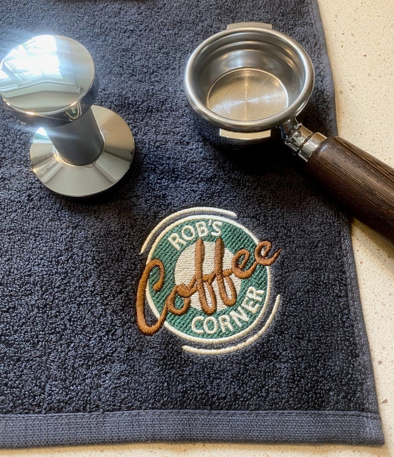 May include: A dark gray towel with a green and white embroidered logo that says "Rob's Coffee Corner". The logo has a brown outline and features a stylized image of a coffee cup. A silver portafilter and tamper are also on the towel.