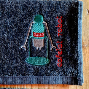May include: A dark blue hand towel with an embroidered image of a coffee robot with the text "coffeelat robot" in red.