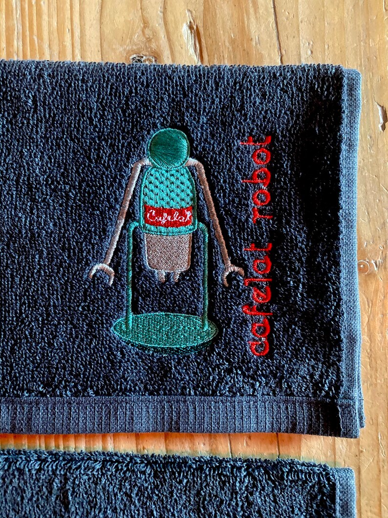 May include: A dark blue hand towel with an embroidered image of a coffee robot with the text "coffeelat robot" in red.