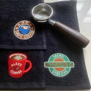 May include: Three black towels featuring embroidered coffee-themed designs. One shows a blue cup with 'DAD'S COFFEE', another a red mug with 'ALEX'S COFFEE', and the third a green logo with 'JACK'S COFFEE STATION'. A metal portafilter with a wooden handle rests on top.