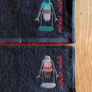 May include: Two black hand towels with embroidered images of a robot holding a coffee maker. The text "coffeelot robot" is embroidered on the towels in red.