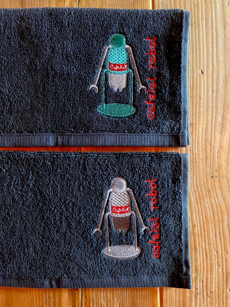 May include: Two black hand towels with embroidered images of a robot holding a coffee maker. The text "coffeelot robot" is embroidered on the towels in red.
