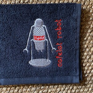 May include: A dark gray hand towel with a white embroidered image of a robot holding a coffee cup. The text "cafe robot" is embroidered in red on the towel.