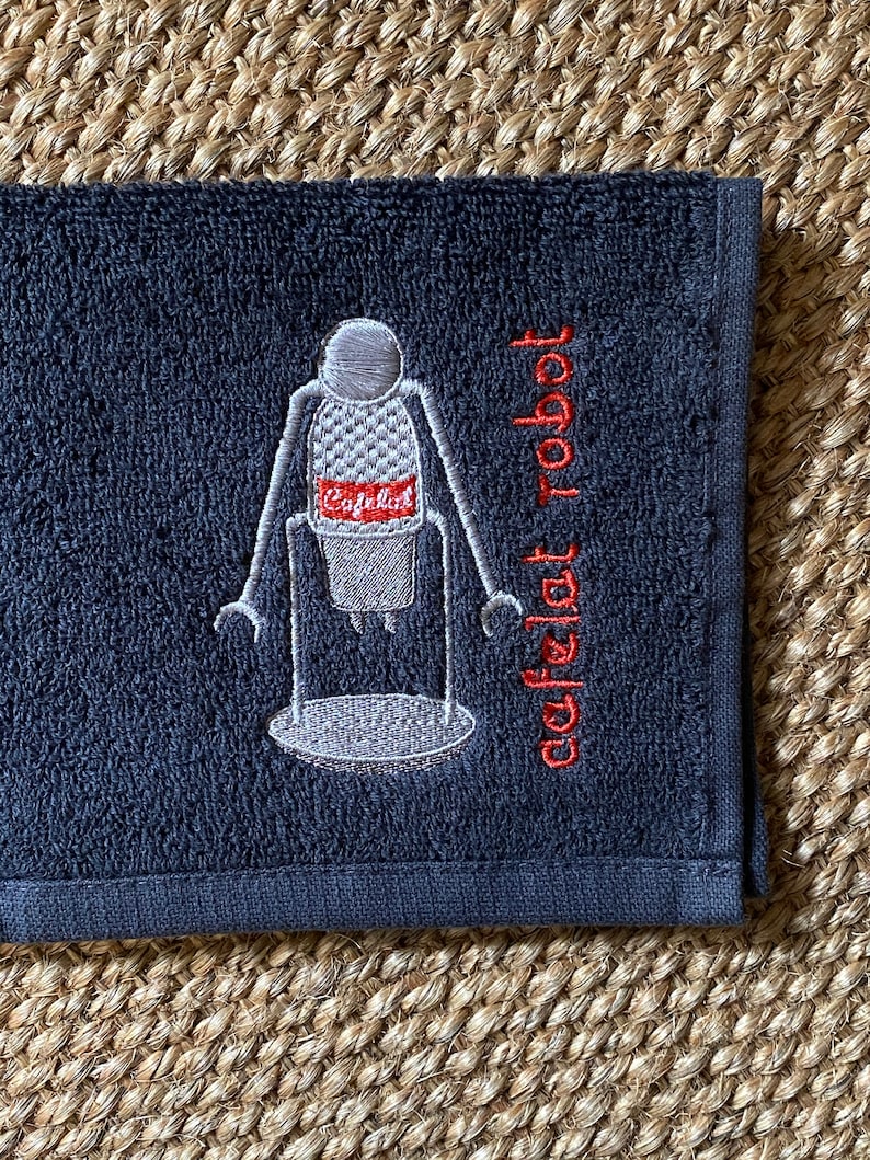May include: A dark gray hand towel with a white embroidered image of a robot holding a coffee cup. The text "cafe robot" is embroidered in red on the towel.