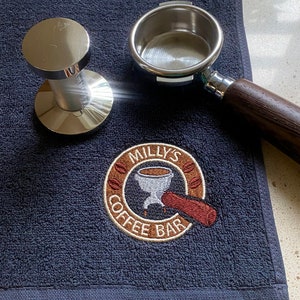 May include: A dark navy blue textured towel featuring an embroidered circular logo that reads MILLY'S COFFEE BAR with a graphic of a portafilter. A silver metal coffee tamper and a metal portafilter with a dark wood handle rest on the towel.