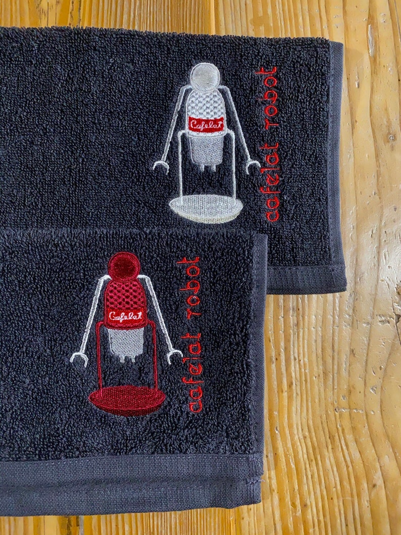 May include: Two dark gray hand towels with embroidered images of a robot holding a coffee maker. The text "Cafelat Robot" is embroidered on each towel.