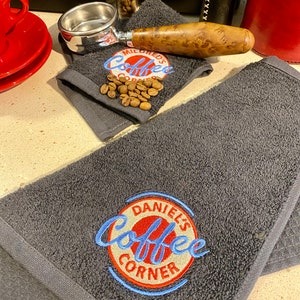 May include: Two dark gray towels with embroidered logos. One towel has the text "Mildred's Coffee Corner" in white with a red circle around it. The other towel has the text "Daniel's Coffee Corner" in blue with a red circle around it.
