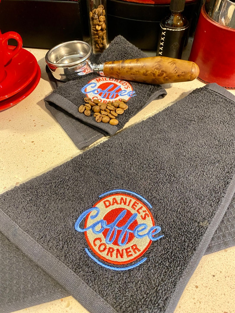 May include: Two dark gray towels with embroidered logos. One towel has the text "Mildred's Coffee Corner" in white with a red circle around it. The other towel has the text "Daniel's Coffee Corner" in blue with a red circle around it.