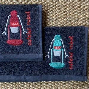 May include: Two dark blue hand towels with embroidered images of a robot holding a coffee cup. The text "cafelat robot" is embroidered on each towel in red.