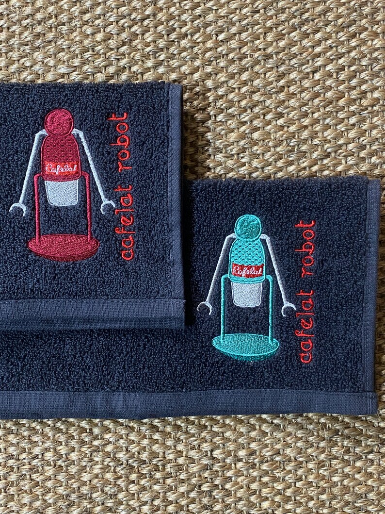May include: Two dark blue hand towels with embroidered images of a robot holding a coffee cup. The text "cafelat robot" is embroidered on each towel in red.