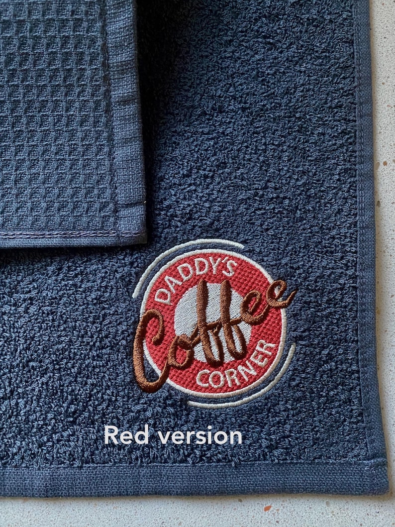 May include: A dark blue kitchen towel with a red and white embroidered logo that says "Daddy's Coffee Corner".
