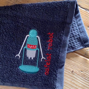 May include: A dark gray kitchen towel with an embroidered robot design. The robot is blue and green with red accents. The text "dafelat robot" is embroidered in red below the robot.