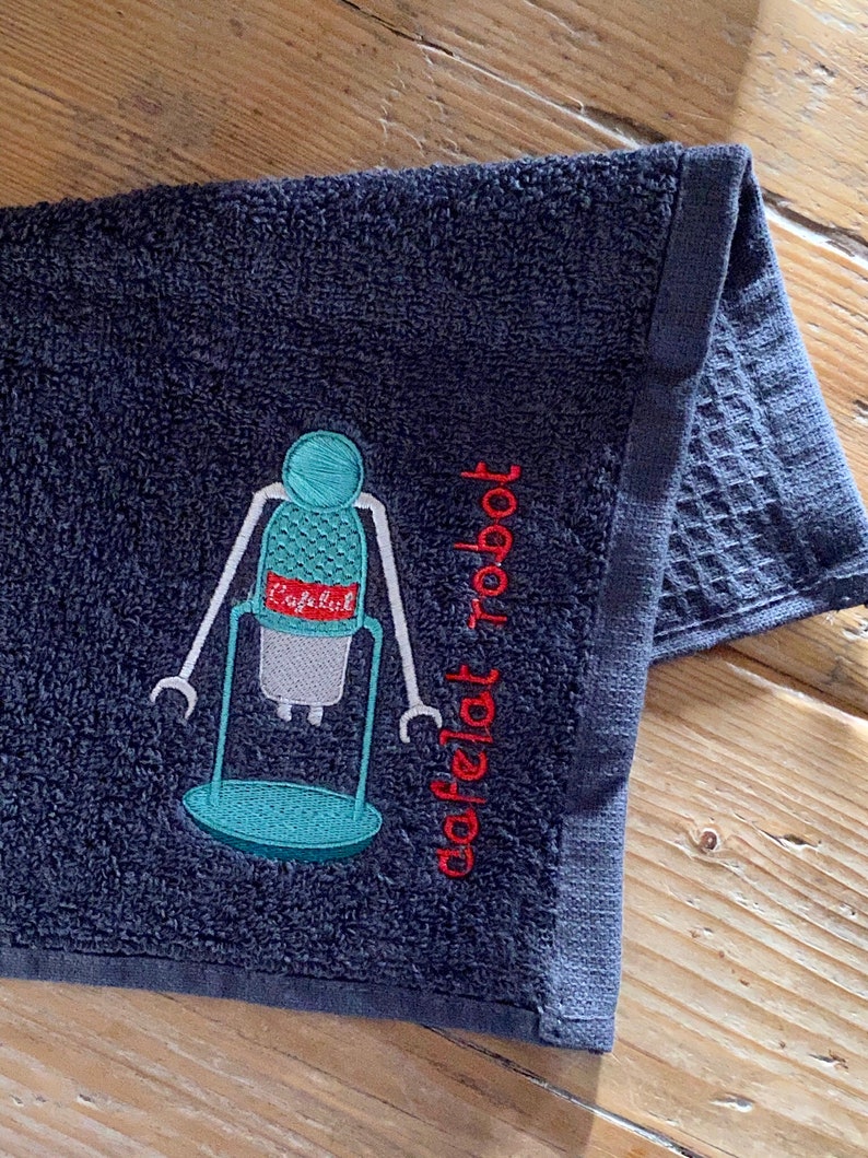 May include: A dark gray kitchen towel with an embroidered robot design. The robot is blue and green with red accents. The text "dafelat robot" is embroidered in red below the robot.