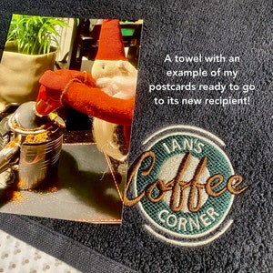 May include: A dark blue towel with a green and brown embroidered logo that says "Ian's Coffee Corner". The towel also has a postcard attached to it with a picture of a gnome pouring coffee grounds into a coffee pot.