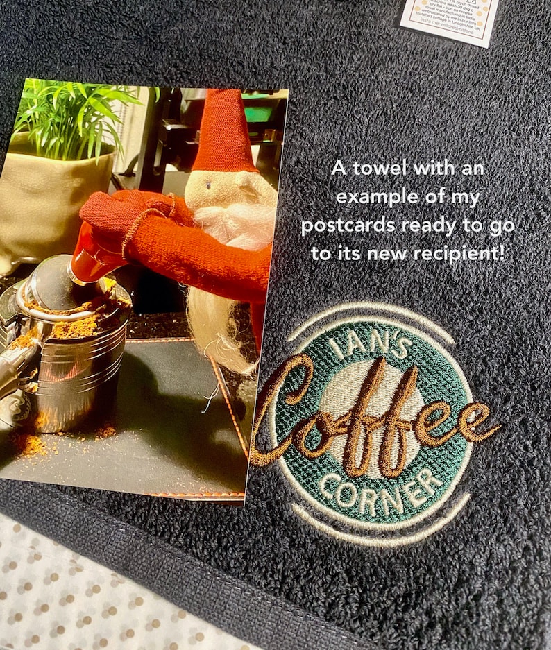 May include: A dark blue towel with a green and brown embroidered logo that says "Ian's Coffee Corner". The towel also has a postcard attached to it with a picture of a gnome pouring coffee grounds into a coffee pot.