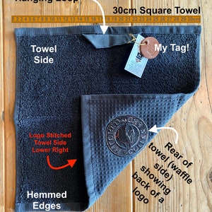 May include: A black waffle weave kitchen towel with a stitched logo in the lower right corner. The towel is 30 inches square and has a hanging loop. The towel is hemmed on all edges. The towel is shown folded in half with the back side of the towel visible. The towel has a brown leather tag with the text "My Tag!" on it.