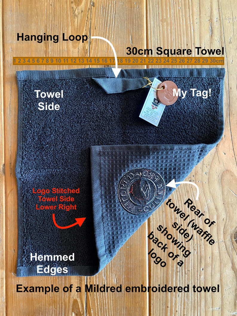 May include: A black waffle weave kitchen towel with a stitched logo in the lower right corner. The towel is 30 inches square and has a hanging loop. The towel is hemmed on all edges. The towel is shown folded in half with the back side of the towel visible. The towel has a brown leather tag with the text "My Tag!" on it.