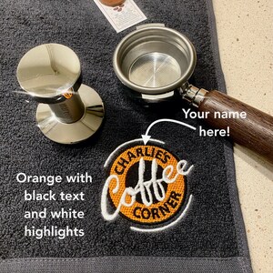 May include: A black coffee towel with an orange embroidered logo that says "Charlie's Coffee Corner". The towel also has a space for a name to be embroidered. A silver portafilter and tamper are also in the image.