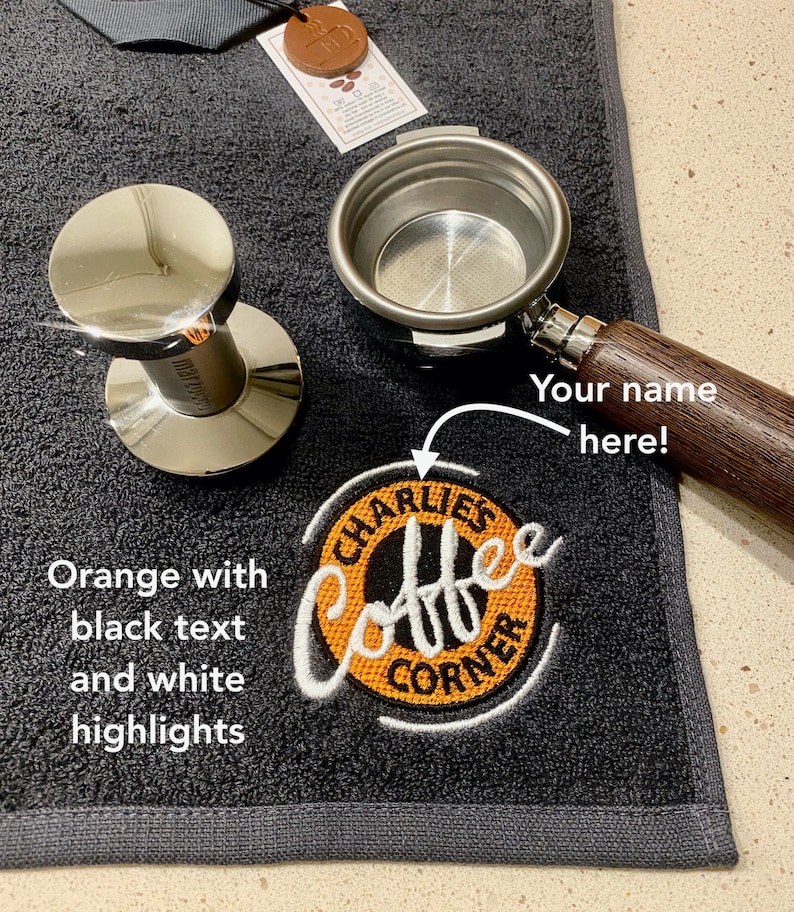 May include: A black coffee towel with an orange embroidered logo that says "Charlie's Coffee Corner". The towel also has a space for a name to be embroidered. A silver portafilter and tamper are also in the image.