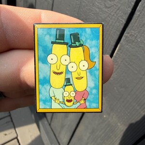 以下が含まれることがあります： 3人の黄色い円筒形のキャラクターの家族を描いた漫画風のイラストが特徴の長方形のエナメルピン。各キャラクターは小さな黒いシルクハットをかぶっています。背景は白い雲が浮かぶ青い空で、黄色い枠で囲まれています。
