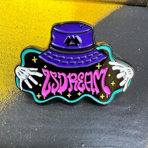 May include: A purple bucket hat pin with the text "LSDREAM" in pink, outlined in black. The pin features white skeletal hands and yellow star accents on a black background with a teal border.