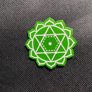May include: A green and white enamel pin featuring the Anahata chakra symbol. The pin has a floral border and geometric design, with a central dot. The pin is a vibrant green color with white accents.