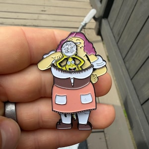 以下が含まれることがあります： 紫色の髪で茶色とピンクのドレスを着た漫画のキャラクターが描かれた金属製のエナメルピン。キャラクターの胸元から現れた小さな黄色い生き物の上に、「SCHMECKLES 25」と書かれた円形のコインを掲げているデザイン。