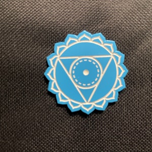 May include: A light blue enamel pin featuring a stylized design with a central circle, a downward-pointing triangle, and a floral border. The pin has white accents and is set against a dark background.