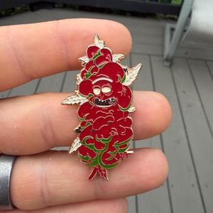 Puede incluir: Un pin de esmalte y metal dorado que presenta un rostro de personaje de dibujos animados estilizado en rojo y verde, integrado en un grupo de hojas. El diseño incluye una expresión de ojos muy abiertos con una boca y dientes marcados.