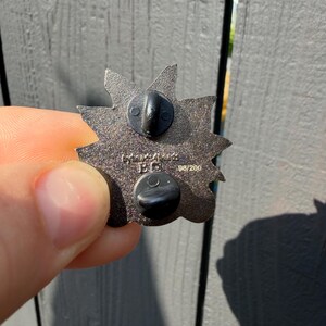 May include: A dark metallic star-shaped enamel pin back featuring two black rubber clutch fasteners. The metal surface has a textured finish with the embossed text "98/200" and additional markings visible on the centre of the pin's reverse side.