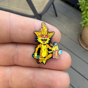 以下が含まれることがあります： 赤い目、広い笑顔、尖った耳を持つ漫画のキャラクターが描かれた黄色のエナメルピン。キャラクターはあぐらをかいて座り、片手に茶色のガラス製水パイプ、もう片方の手に小さな青い道具を持っています。