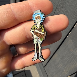 May include: A metal enamel pin featuring a cartoon character with light blue hair, wearing orange-tinted glasses, a yellow floral print shirt, and white shorts. The character holds a brown rectangular object with a silver handle and green accents.