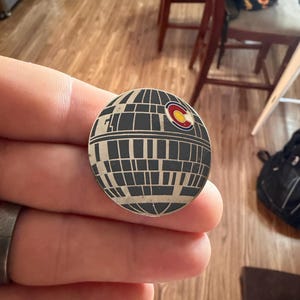 Puede incluir: Un pin redondo plateado con un diseño detallado de la Estrella de la Muerte en negro y gris. La bandera del estado de Colorado está en el lado derecho del pin. El pin está en la mano de una persona.