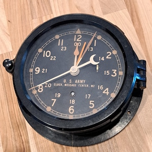 May include: A black U.S. Army clock with a round face and a black bezel. The clock face has white and tan numerals and hands. The text "U.S. ARMY CLOCK, MESSAGE CENTER, M2" is printed on the face.