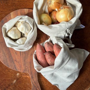 Pode incluir: Tr&ecirc;s sacos de tecido reutiliz&aacute;veis ​​cheios de produtos frescos. O primeiro saco cont&eacute;m alho, o segundo cebolas e o terceiro batatas doces.