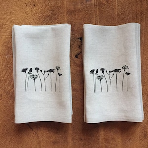 May include: Two linen kitchen towels with a black floral print. The print features a row of stylized flowers and stems.