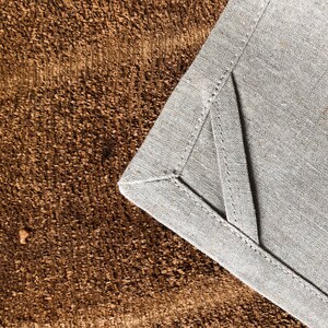 May include: Close-up of a gray linen napkin with a folded corner, showing the stitching detail. The napkin is on a brown textured surface.