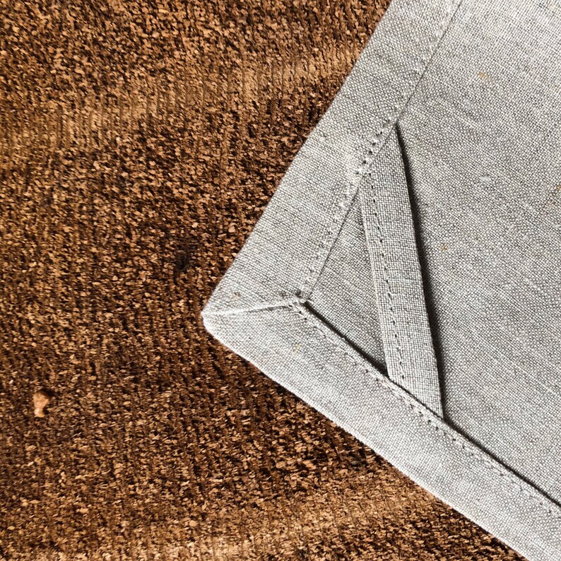 May include: Close-up of a gray linen napkin with a folded corner, showing the stitching detail. The napkin is on a brown textured surface.