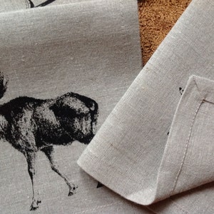 May include: Two linen placemats with a black moose design. The placemats are folded and show the hemmed edges.