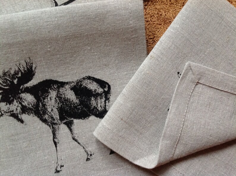 May include: Two linen placemats with a black moose design. The placemats are folded and show the hemmed edges.