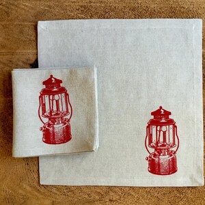 May include: Two linen napkins with a red vintage lantern design. The napkins are folded and placed on a wooden surface.