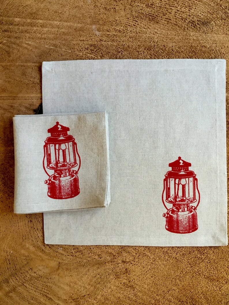 May include: Two linen napkins with a red vintage lantern design. The napkins are folded and placed on a wooden surface.