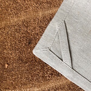 May include: A close-up of a gray linen napkin with a stitched edge. The napkin is folded in a triangle shape and is lying on a brown textured surface.