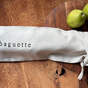 May include: A beige linen bag with the word "baguette" printed on it. The bag is lying on a wooden surface with two green pears and a sprig of rosemary.