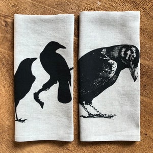 以下が含まれることがあります： 黒いカラスのシルエットがプリントされたリネン製のティータオル2枚。1枚は、枝にとまっているカラスと飛んでいるカラスの2羽が描かれている。もう1枚は、頭を横に傾けたカラスが1羽描かれている。