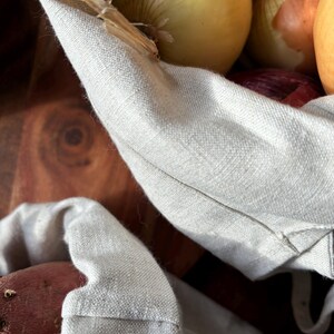 Pode incluir: Um close-up de uma bolsa de linho branca cheia de cebolas e batatas vermelhas. A bolsa est&aacute; dobrada sobre o topo dos vegetais.