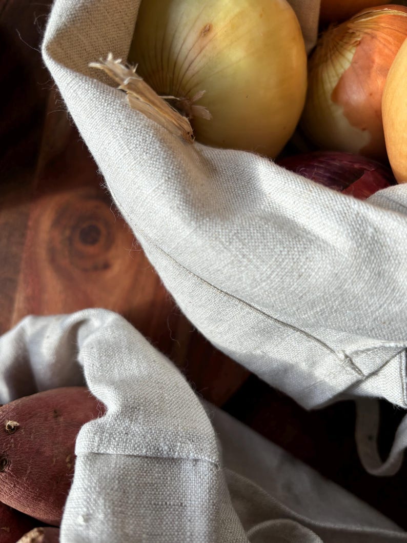 Pode incluir: Um close-up de uma bolsa de linho branca cheia de cebolas e batatas vermelhas. A bolsa est&aacute; dobrada sobre o topo dos vegetais.